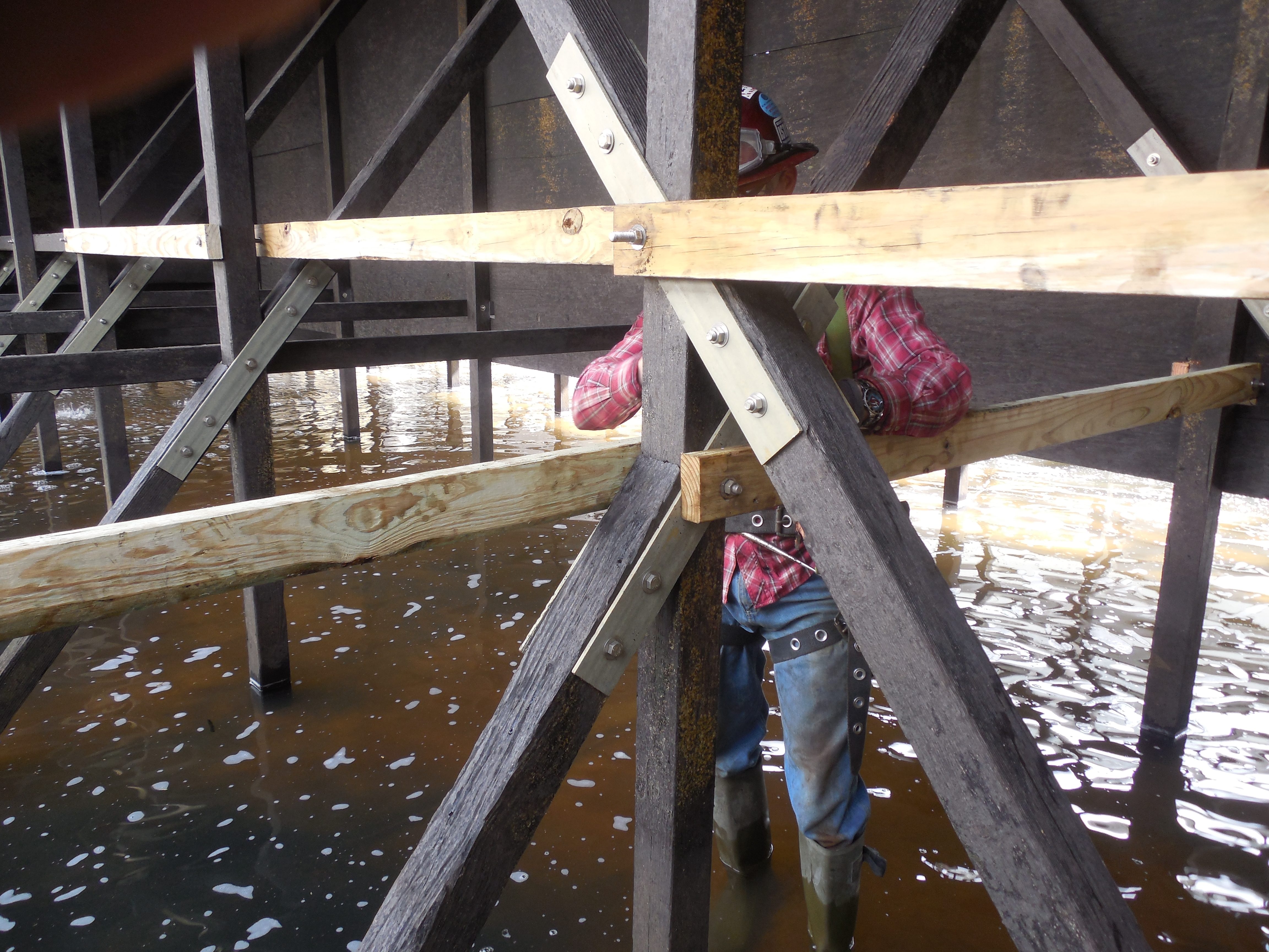 New pressure-treated lumber bracing and galvanized connector plates bolted into aged timber framing inside a flooded cooling tower basin.